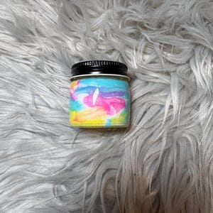 May include: A small glass jar with a black lid, filled with colorful, swirled wax in shades of pink, blue, yellow, and white. The jar sits on a fluffy, gray surface.
