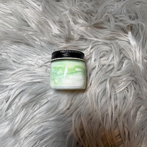 May include: A small, square jar with a black lid. The jar contains a green and white swirled substance, with a white leaf design on the front. The jar is set against a fluffy, gray background.