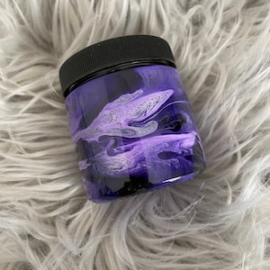 May include: A small glass jar with a black lid. The jar is filled with a purple and white liquid with swirls and marbling.
