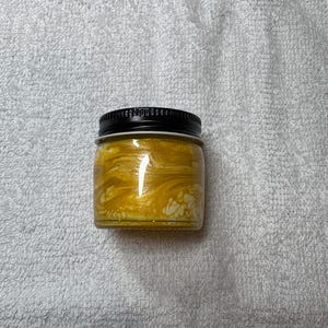 May include: A small glass jar with a black lid filled with a yellow, swirled substance. The jar is sitting on a white, textured surface. The contents of the jar appear to be a cosmetic product.