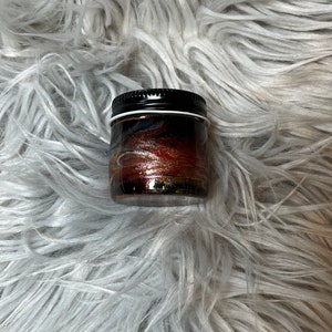 May include: A small glass jar with a black lid. The jar is filled with a glittery, multi-colored liquid that resembles a galaxy.