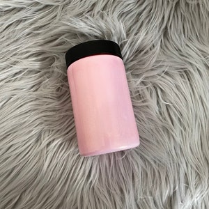 May include: A cylindrical, light pink container with a black lid. The container is set against a backdrop of long, gray faux fur. The container appears to be made of plastic or a similar material.