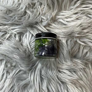 May include: A small glass jar with a black lid filled with dried herbs and flowers. The jar is sitting on a fluffy gray surface.