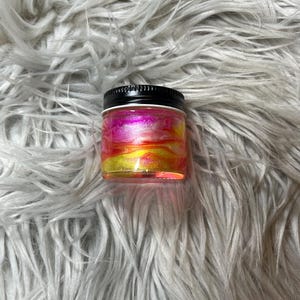 May include: A small glass jar with a black lid filled with a glittery pink, yellow, and orange liquid.