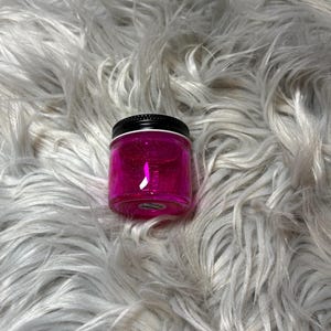 May include: A small, clear jar filled with bright pink glitter gel. The jar has a black lid and a small, round label. The jar is set against a backdrop of fluffy, white faux fur.