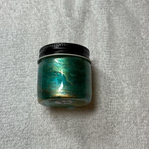 May include: A small, clear glass jar with a black lid. The jar is filled with a shimmering, emerald green substance with gold accents. The jar is sitting on a white, textured surface.