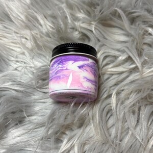 May include: A small, round jar with a black lid. The jar's exterior features a swirling pattern of purple, pink, and white. The jar is sitting on a fluffy, gray surface.
