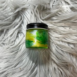 May include: A small glass jar with a black lid filled with green and yellow glitter. The glitter is swirled together to create a marbled effect.