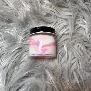 May include: A small glass jar with a black lid, filled with a pink and white swirled substance. The jar is set against a backdrop of fluffy, gray faux fur.