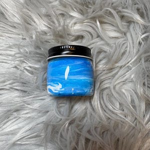 May include: A small, round jar with a black lid. The jar contains a blue and white swirled substance, possibly a candle or cosmetic product. The jar is set against a fluffy, gray background.
