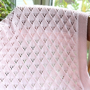 Può includere: Una copertina per neonati rosa all'uncinetto con un motivo a rombi. La copertina è appoggiata su una sedia di legno.