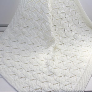 May include: A white crocheted baby blanket with a delicate, lacy pattern. The blanket is folded and draped over a white surface.