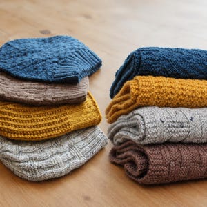 May include: A collection of folded knit hats and scarves in various colors including blue, brown, mustard yellow, and light gray. The items feature diverse textured patterns such as ribbing, cable knits, and basketweave designs, stacked neatly.