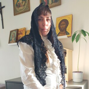 May include: A woman wearing a white blouse and a black lace mantilla. She is standing in front of a wall with religious icons.