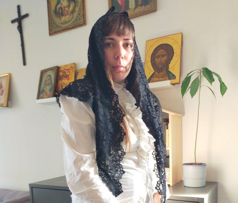 May include: A woman wearing a white blouse and a black lace mantilla. She is standing in front of a wall with religious icons.