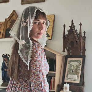 May include: A delicate white veil with floral embroidery and a scalloped lace trim. The veil is worn over the head and shoulders. The person is wearing a floral print dress.