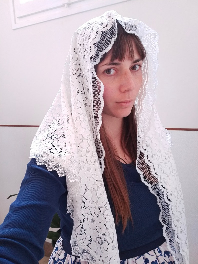 May include: A white lace veil with a floral pattern. The veil is worn over a person's head and shoulders.