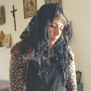 May include: A black lace veil with floral detailing. The veil is draped over the head and shoulders. The person is wearing a black corduroy overall dress and a floral print top.