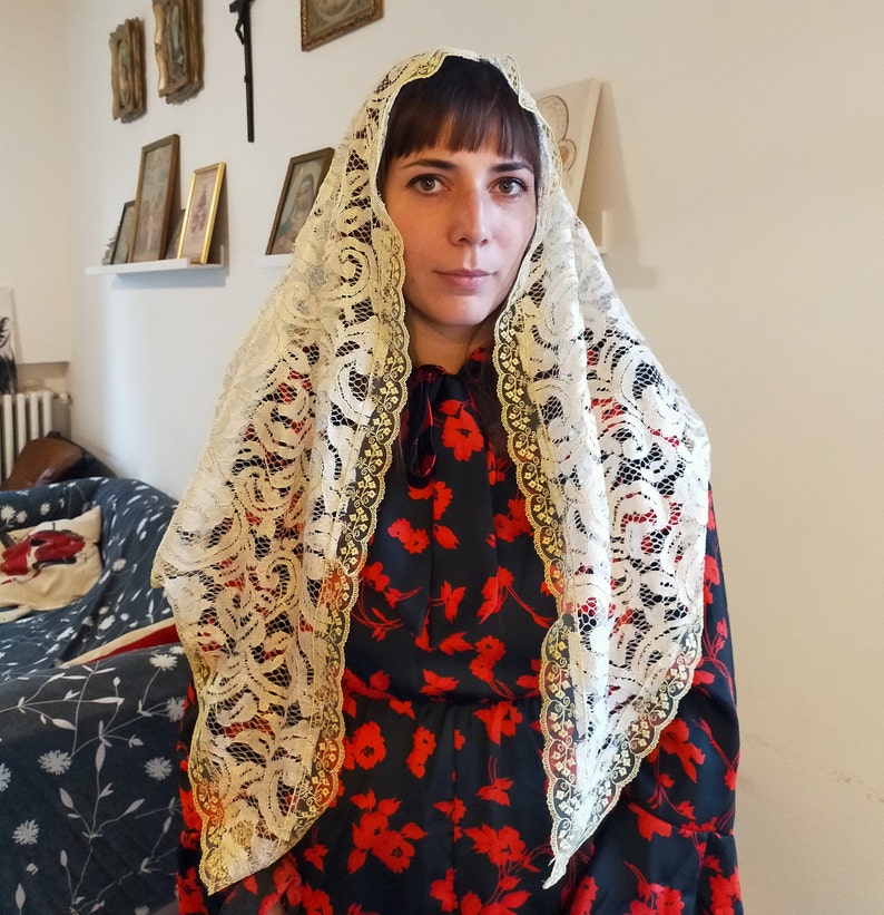 May include: A woman wearing a white lace veil over a black dress with red floral print. The veil is a traditional head covering.
