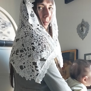 May include: A white lace head covering with a floral design. The lace is draped over the head and shoulders. The person is wearing a gray blazer.