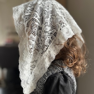 Puede incluir: Un velo de encaje blanco, drapeado sobre la cabeza y los hombros. El delicado encaje presenta un patrón floral. La persona lleva una parte superior negra y gris.