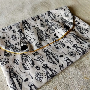 Può includere: Una pochette in tessuto bianco con un bordo giallo e chiusure a bottone nero. La pochette è decorata con un motivo ripetitivo di illustrazioni in bianco e nero di una figura religiosa con corona e tunica.