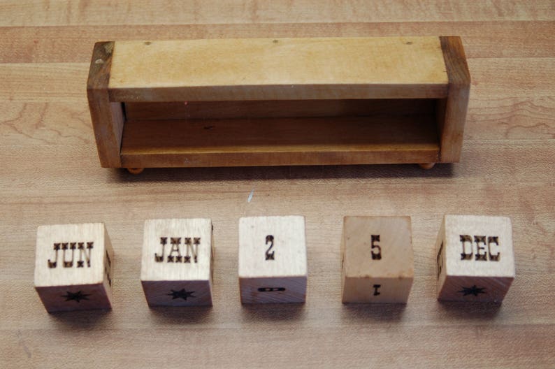 Puede incluir: Calendario perpetuo de madera clara. Incluye una estanter&iacute;a de madera y cinco bloques de madera. Los bloques muestran los meses de junio, enero y diciembre, junto con los n&uacute;meros 2 y 5. Cada bloque de mes tiene un dise&ntilde;o de estrella.
