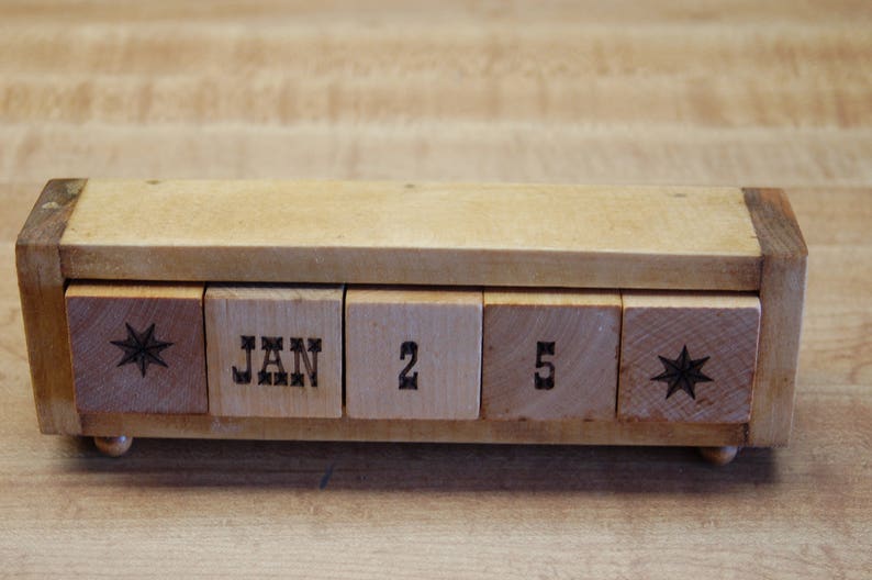 Puede incluir: Calendario perpetuo de madera que muestra "JAN 2 5". El calendario tiene un marco rectangular de madera y peque&ntilde;os pies. Los bloques son de color marr&oacute;n claro con texto marr&oacute;n oscuro y dise&ntilde;os de estrellas. Un art&iacute;culo decorativo para un escritorio o estante.