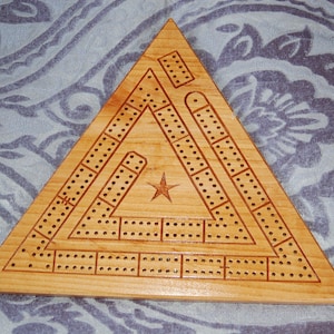 May include: A wooden cribbage board in a triangular shape. The board is made of light-colored wood and features a star in the center. It has multiple rows of holes for pegs, and the design is etched into the wood.