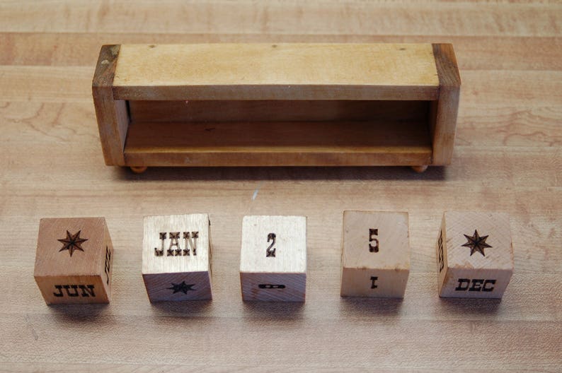 Puede incluir: Calendario perpetuo de madera con acabado marr&oacute;n claro. El calendario incluye una caja de madera y cinco cubos de madera. Los cubos muestran meses, n&uacute;meros y dise&ntilde;os de estrellas. Los meses mostrados son junio, enero y diciembre.