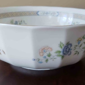 May include: A white porcelain bowl with a faceted, scalloped rim and a gold-trimmed edge. The exterior features a delicate floral pattern with blue, peach, and green flowers and leaves, complemented by a geometric band along the inner rim.