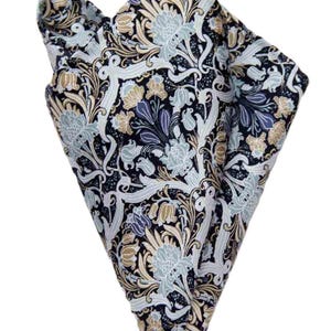 May include: A folded pocket square featuring a dark navy blue background with an intricate floral pattern in shades of light blue, cream, and tan. The design consists of stylized flowers, swirling leaves, and vines covering the entire fabric surface.