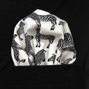 Op de afbeelding: Een wit pochet met een herhalend zwart zebramotief. De stof is gevouwen met zachte, afgeronde plooien en toont meerdere zebra's in verschillende posities over het oppervlak van het materiaal tegen een effen zwarte achtergrond.