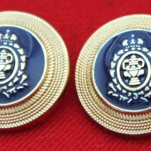 May include: Two circular buttons featuring a deep blue center with a raised gold-tone emblem of a crown above a central vessel surrounded by a wreath. The outer edge of each button has a textured gold-tone border with a fine beaded pattern.
