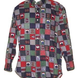 Puede incluir: Camisa abotonada de manga larga con un diseño de retazos en cuadrados rojos, verdes, azul marino y grises. La camisa presenta los números "96" y "19", junto con la letra "C" y otros elementos gráficos. La camisa se muestra en un maniquí blanco.