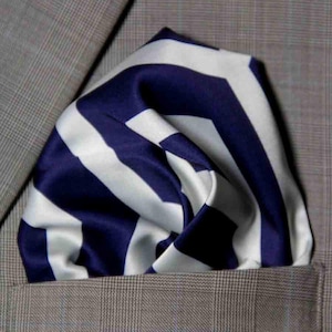 May include: A pocket square featuring a bold, navy blue and white chevron pattern. The fabric has a smooth, satin-like finish and is folded into a puff style, tucked neatly into the breast pocket of a light brown, plaid-patterned suit jacket.