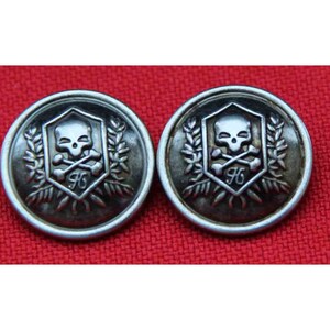 May include: Two circular silver-toned metal buttons featuring a raised emblem of a skull and crossbones centered within a shield. The shield is flanked by leafy branches and includes a small letter H at the base of the skull design.