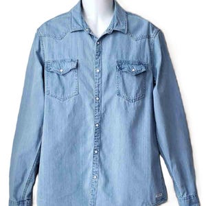 Puede incluir: Camisa vaquera azul claro con mangas largas, dos bolsillos en el pecho y cierre de botones. La camisa tiene un cuello cl&aacute;sico y detalles de estilo occidental. La tela parece ser un denim de peso medio.