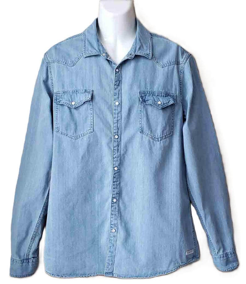 Puede incluir: Camisa vaquera azul claro con mangas largas, dos bolsillos en el pecho y cierre de botones. La camisa tiene un cuello cl&aacute;sico y detalles de estilo occidental. La tela parece ser un denim de peso medio.