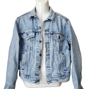 May include: Light blue denim jacket with button closures, two chest pockets, and side pockets. The jacket is displayed on a white mannequin. A small red tag is visible on the left chest pocket.