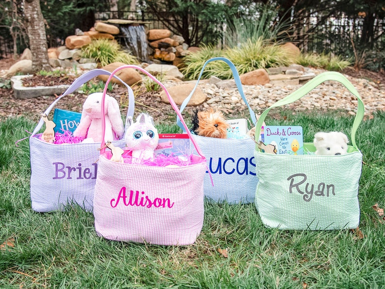 May include: Four personalized Easter baskets with pastel colors and handles. The baskets are filled with stuffed animals, books, and Easter grass. The names "Briel", "Allison", "Lucas", and "Ryan" are embroidered on the baskets.