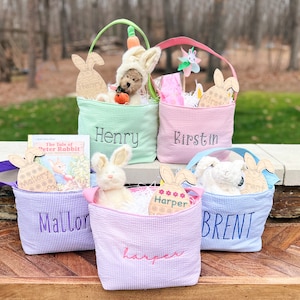 Op de afbeelding: Vier gepersonaliseerde paaskorven gevuld met verschillende items, waaronder een knuffelkonijn, een boek en een houten ei. De korven zijn in groen, roze, paars en blauw gingham.