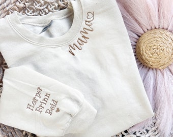 May include: A light beige sweatshirt with the word "mama" embroidered on the neckline. The sweatshirt has a long sleeve and a small heart embroidered on the cuff. The text "Harper Brianna Reid" is embroidered on the cuff.