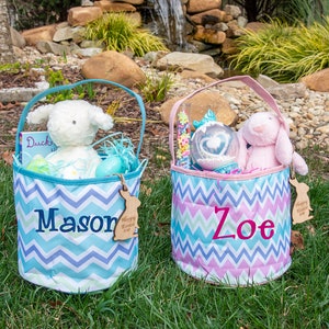 May include: Two Easter baskets with personalized names, Mason and Zoe. The blue basket has a white lamb stuffed animal, a book, and Easter eggs. The pink basket has a pink bunny stuffed animal, a clear plastic globe with colorful confetti, and a pink and white candy cane. Both baskets have a chevron pattern and a wooden bunny tag.