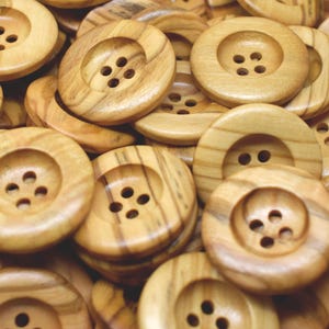 May include: A collection of round, light brown wooden buttons with visible grain patterns. Each button features a recessed centre and four holes for sewing. The buttons are stacked and overlapping, showing a smooth, polished finish throughout.