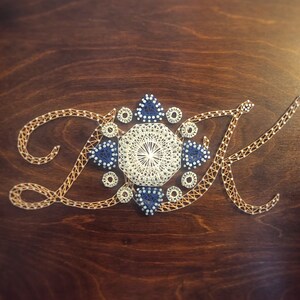 May include: A close-up of a wooden surface with the letters "DK" created using string art. The letters are surrounded by a white and blue geometric design.