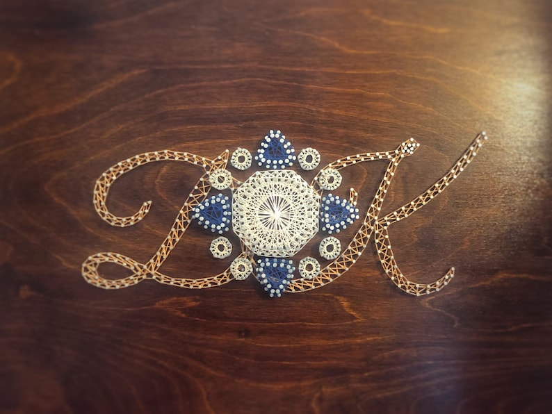 May include: A close-up of a wooden surface with the letters "DK" created using string art. The letters are surrounded by a white and blue geometric design.