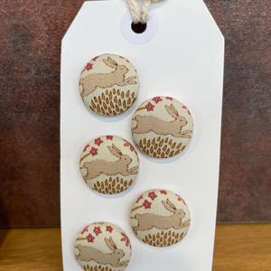 May include: A white tag with five fabric-covered buttons. Each button features a running rabbit design in tan and red on a cream background. The tag has a string loop and the text "Buttonhom" at the bottom.