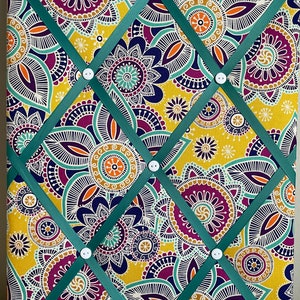 Puede incluir: Un tablero de notas decorativo con un vibrante estampado floral en amarillo, morado, turquesa y naranja. Cintas turquesas se cruzan sobre la superficie, aseguradas por botones blancos. Un lazo de cinta turquesa en la parte superior para colgar.