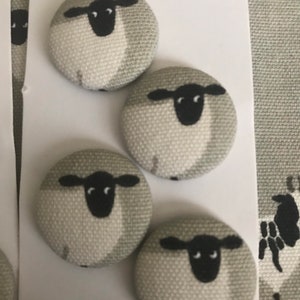 May include: Four round fabric buttons with a sheep design. Each button has a light grey background with a black and white sheep illustration. The buttons are attached to a white tag.